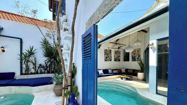 Un área de piscina exterior moderna con una puerta azul, un espacio de descanso con cojines y diversas plantas.