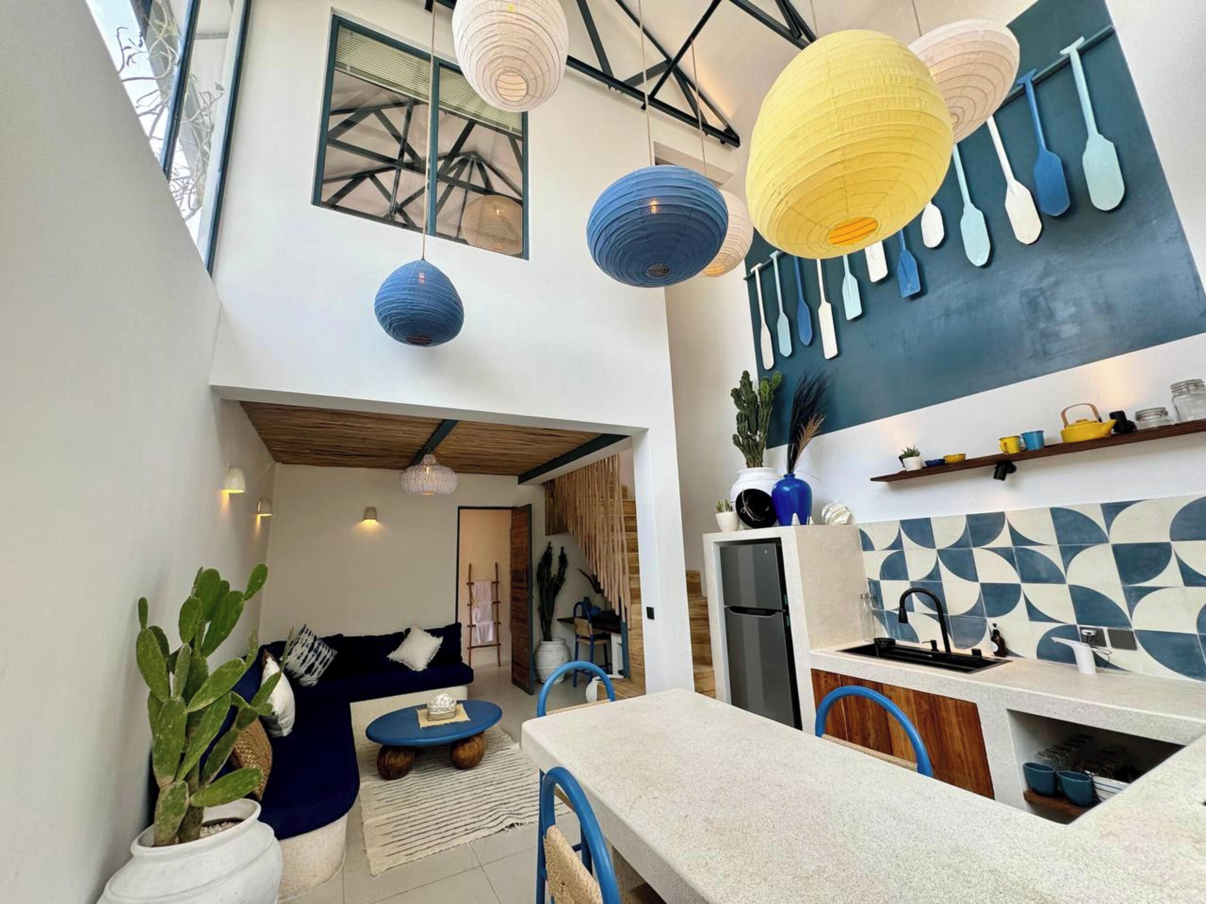 A modern kitchen with a dining area, featuring a unique geometric backsplash, pendant lights, and a variety of kitchenware on shelves.