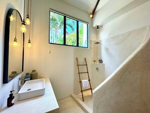 A modern bathroom with a white sink, a mirror, and a shower. There is a window with a view of greenery outside. - Santorini Studio (Canggu)