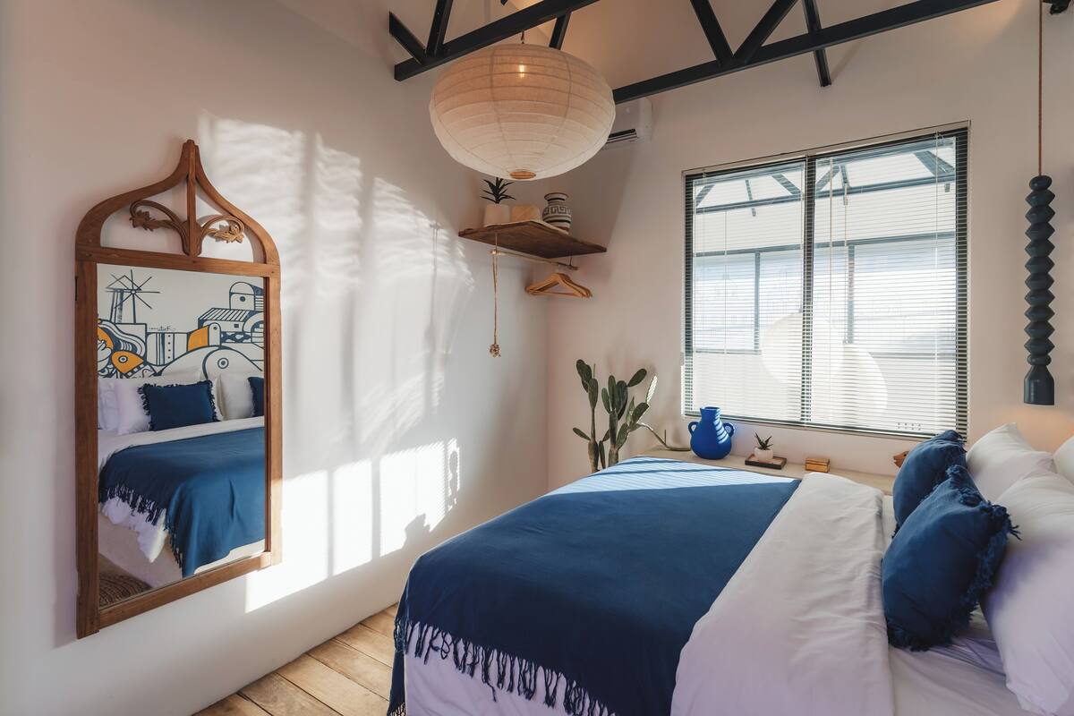 Une chambre à coucher avec un lit, un grand miroir, un luminaire suspendu, une fenêtre avec des stores et une poutre en bois au plafond.