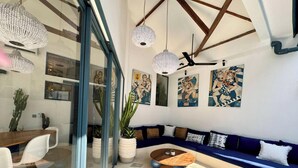 Una sala de estar moderna con un sofá azul, una mesa de centro de madera y un separador de vidrio. Se observan ventiladores de techo, lámparas colgantes y obras de arte en las paredes.