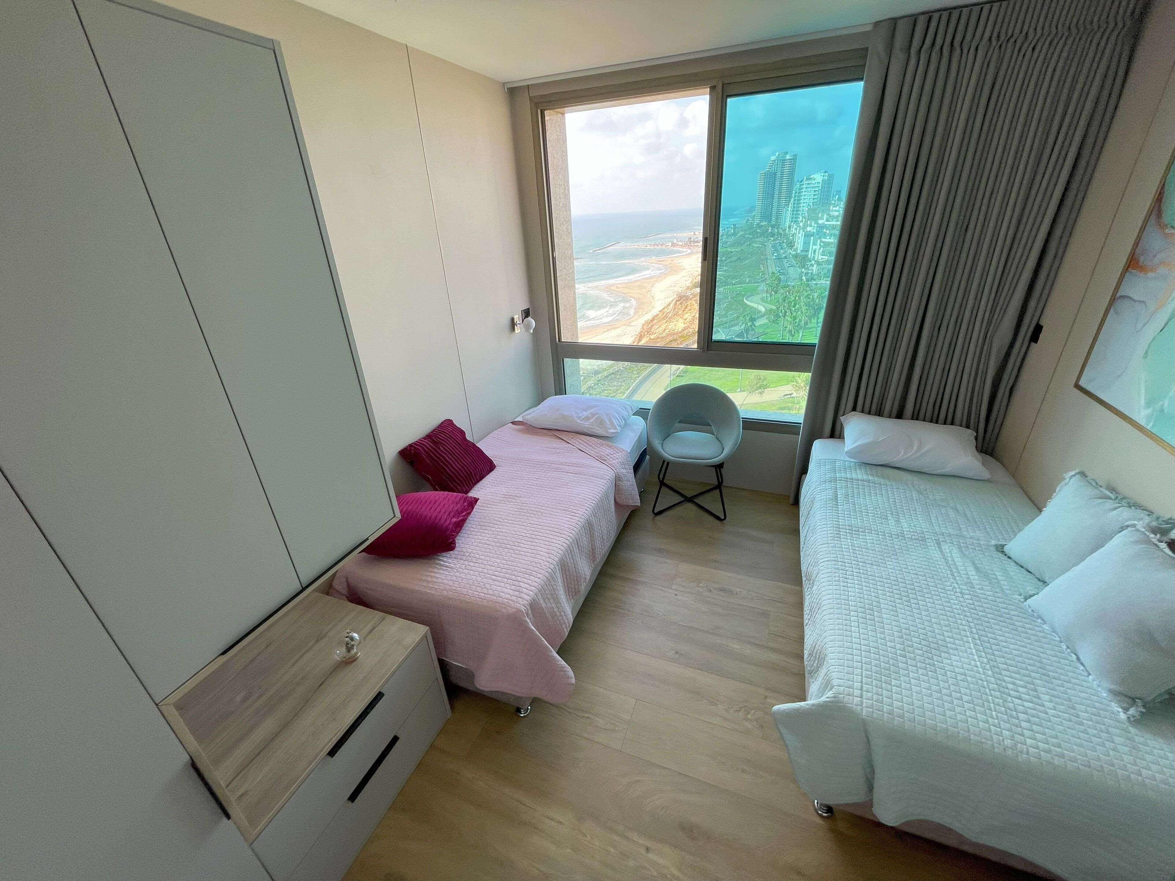 Habitació d'hotel amb dos llits, una cadira, un armari i un finestra gran amb vista a una platja i edificis.