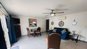 Una sala de estar con un sofá azul, una mesa de comedor con sillas, un ventilador de techo y un cuadro en la pared.