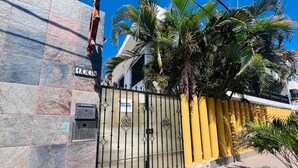 Una entrada protegida con un portón metálico, un muro de azulejos y una pared amarilla con palmeras.
