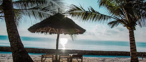 A beach with a thatched umbrella, wooden chairs, and palm trees.