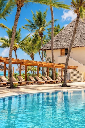 A pool area with lounge chairs and a thatched-roof building. - JAZ Elite Adonia (Uroa)