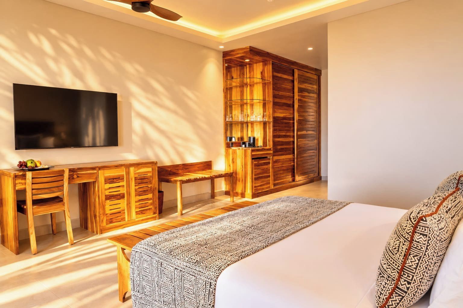 A modern bedroom with a large bed, a desk with chairs, a television, and built-in wooden shelving.