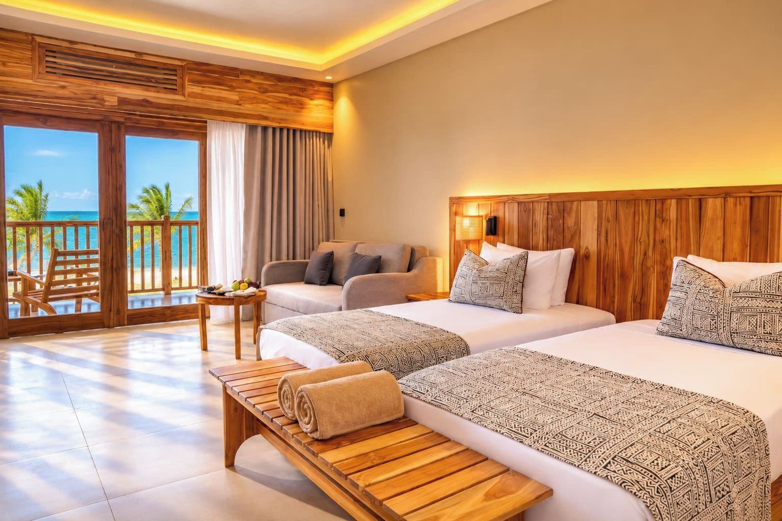Habitación de hotel con dos camas, un sofá, un banco de madera, una mesita y vista al océano.