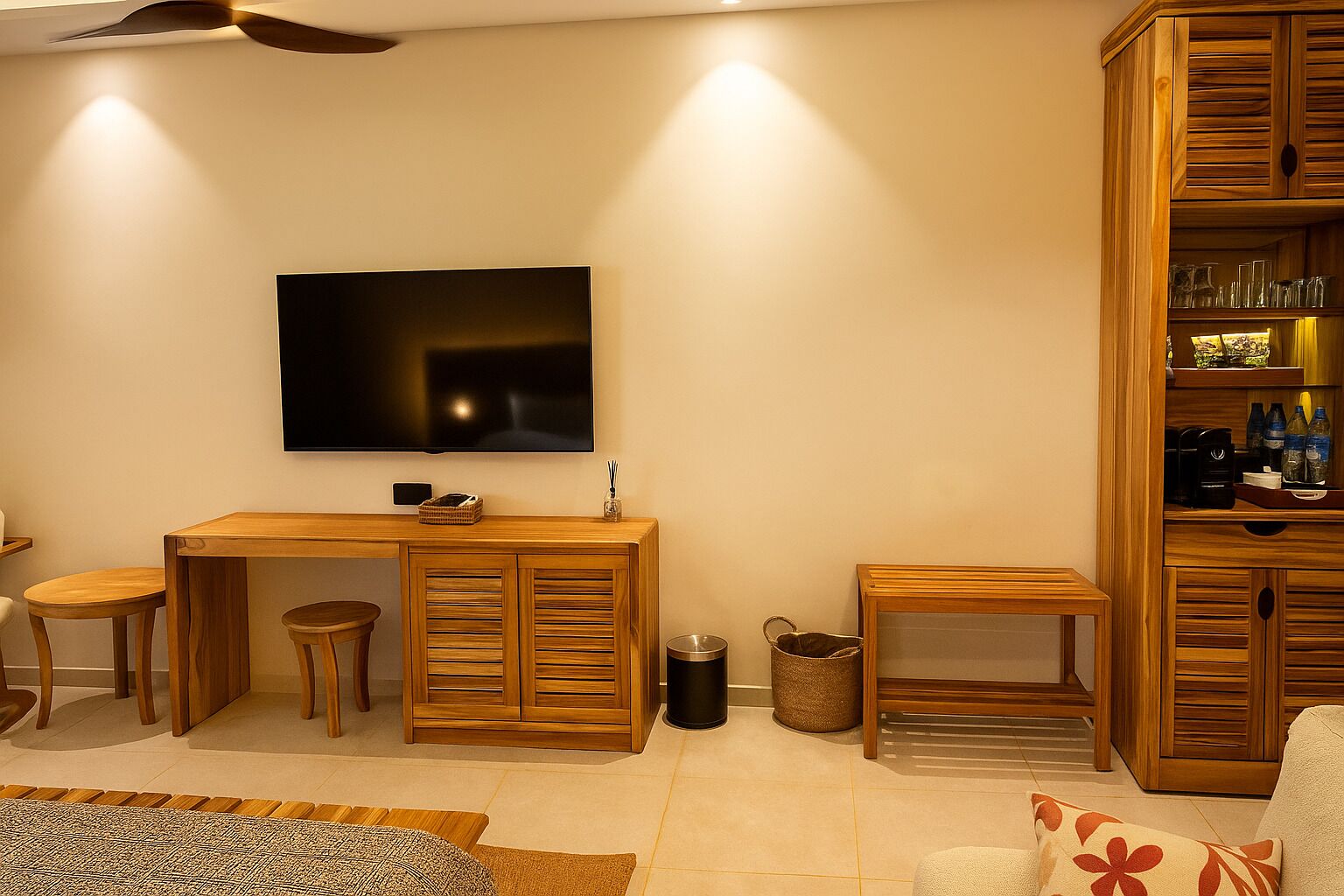 A modern living room with a flat-screen TV mounted on the wall, wooden furniture, a coffee machine, and a small cabinet with glassware.