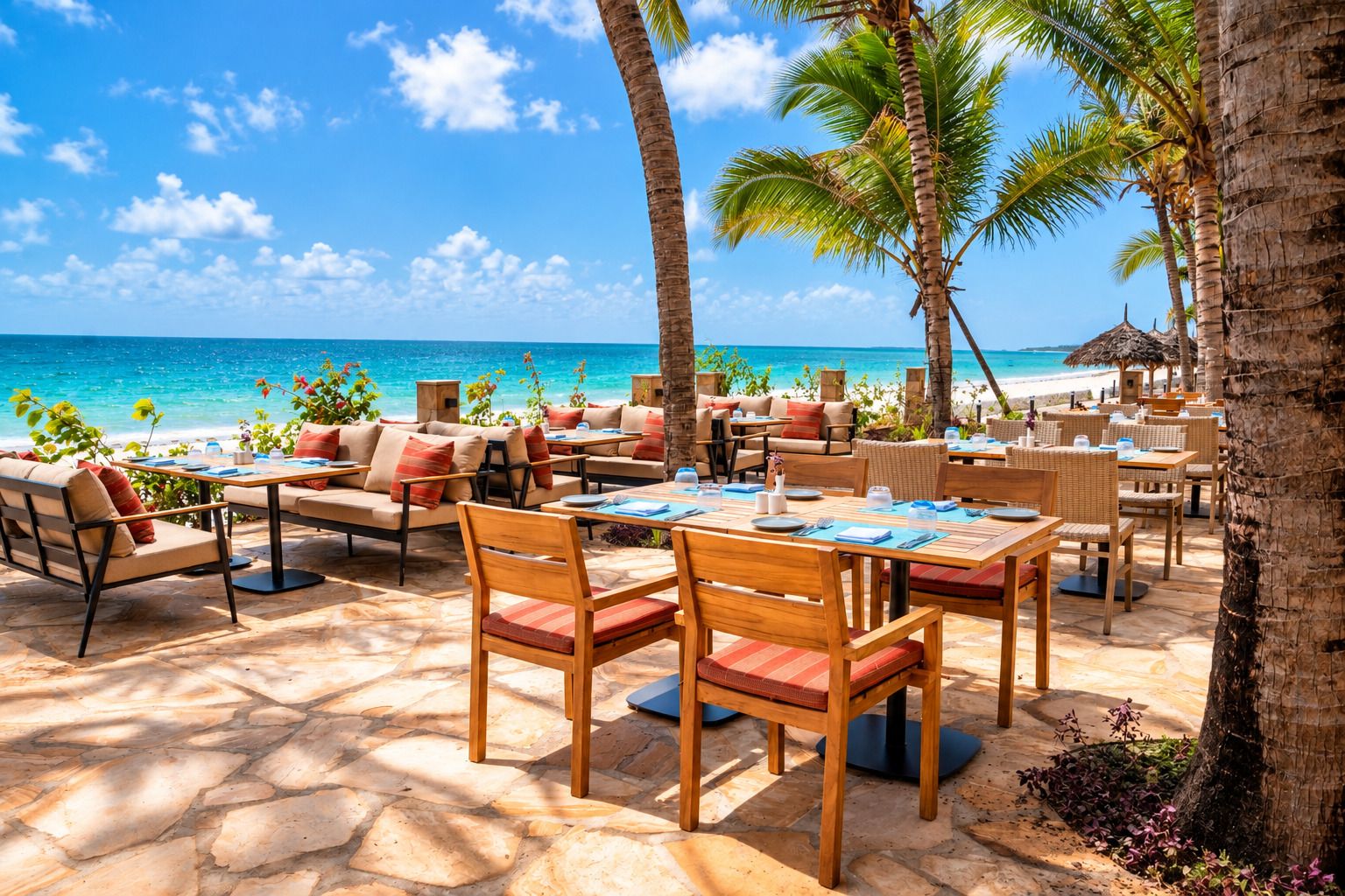 An outdoor dining area with tables and chairs, palm trees, and a view of the ocean.