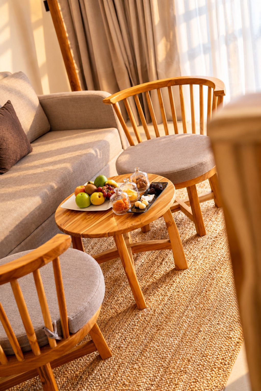 A cozy seating area with a round wooden table set with fruits and nuts, a sofa, and two wooden chairs.