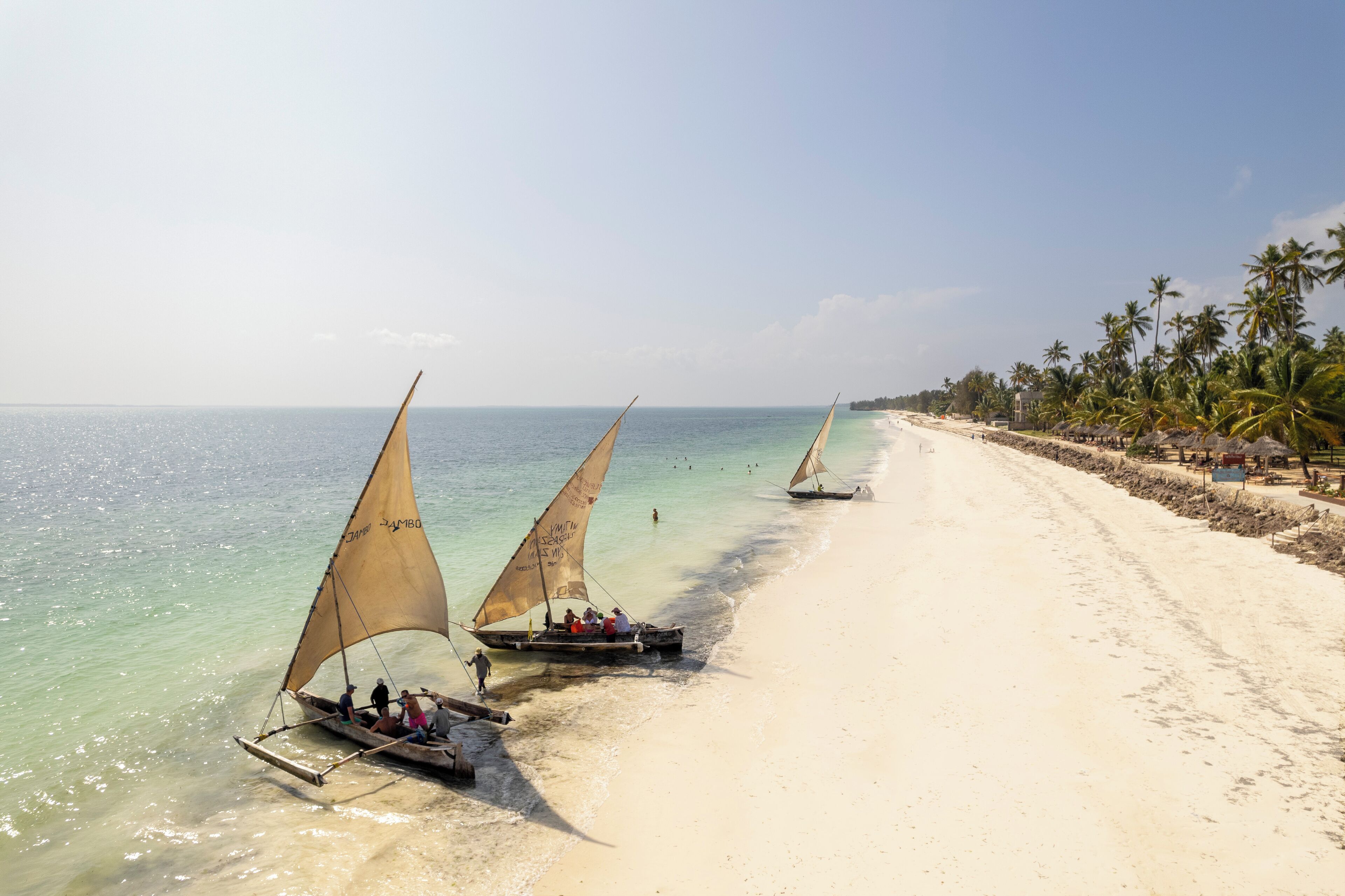 Una playa con agua turquesa cristalina, palmeras y barcos con velas.