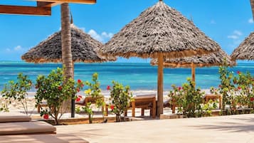 A beachside lounge area with wooden benches and thatched umbrellas, overlooking a clear blue sea.
