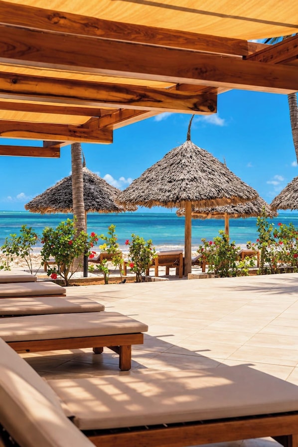 A beachside lounge area with wooden benches and thatched umbrellas, overlooking a clear blue sea. - JAZ Elite Adonia (Uroa)