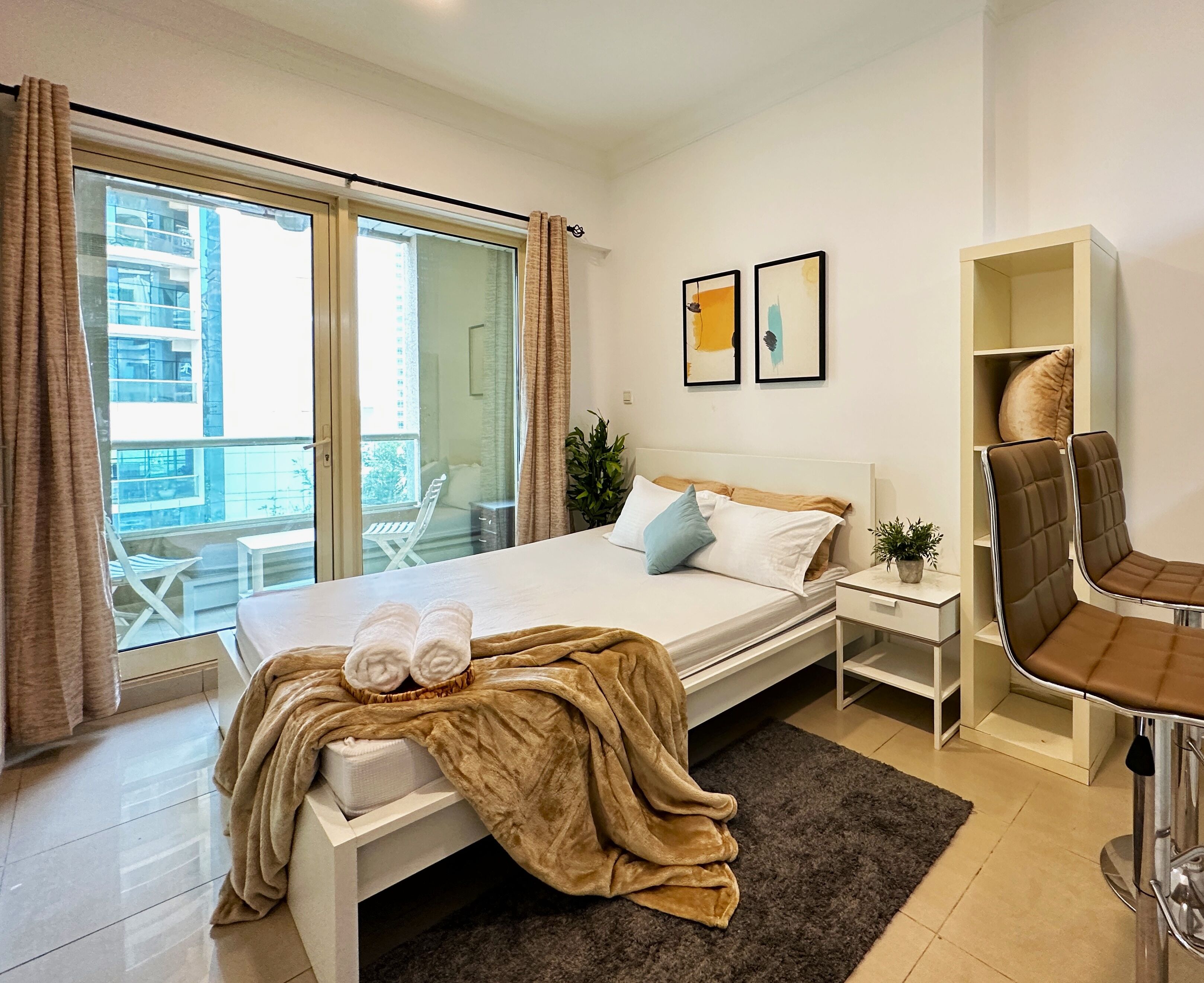 A modern hotel room with a bed, a small bedside table, a chair, a window with curtains, and a view of the outside balcony.