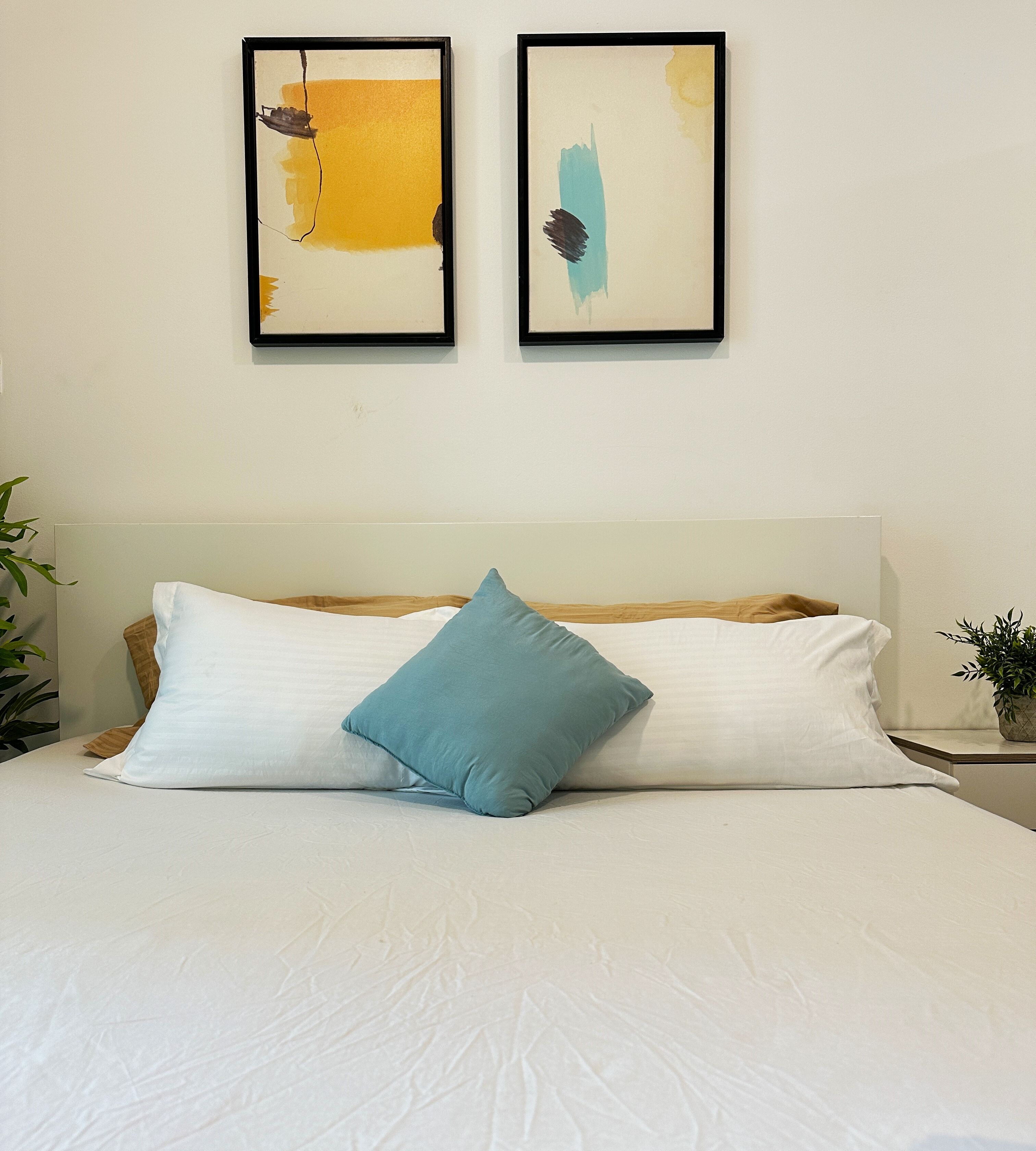 Een bed met witte beddengoed en één smaragdgroen kussen, een houten hoofdbord en twee abstracte schilderijen aan de muur.