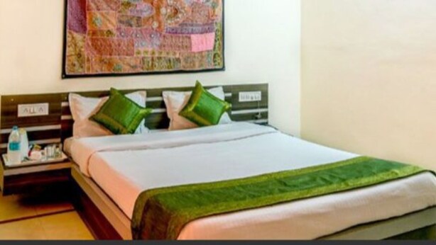 A hotel room with a bed, a nightstand, a wall-mounted quilt, and a painting above the bed. - Golden Home Hotel By GRB (Jaisalmer)