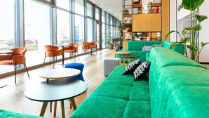 Een moderne lounge met een groene bank, oranje fauteuils, ronde tafels en grote ramen met uitzicht buiten.