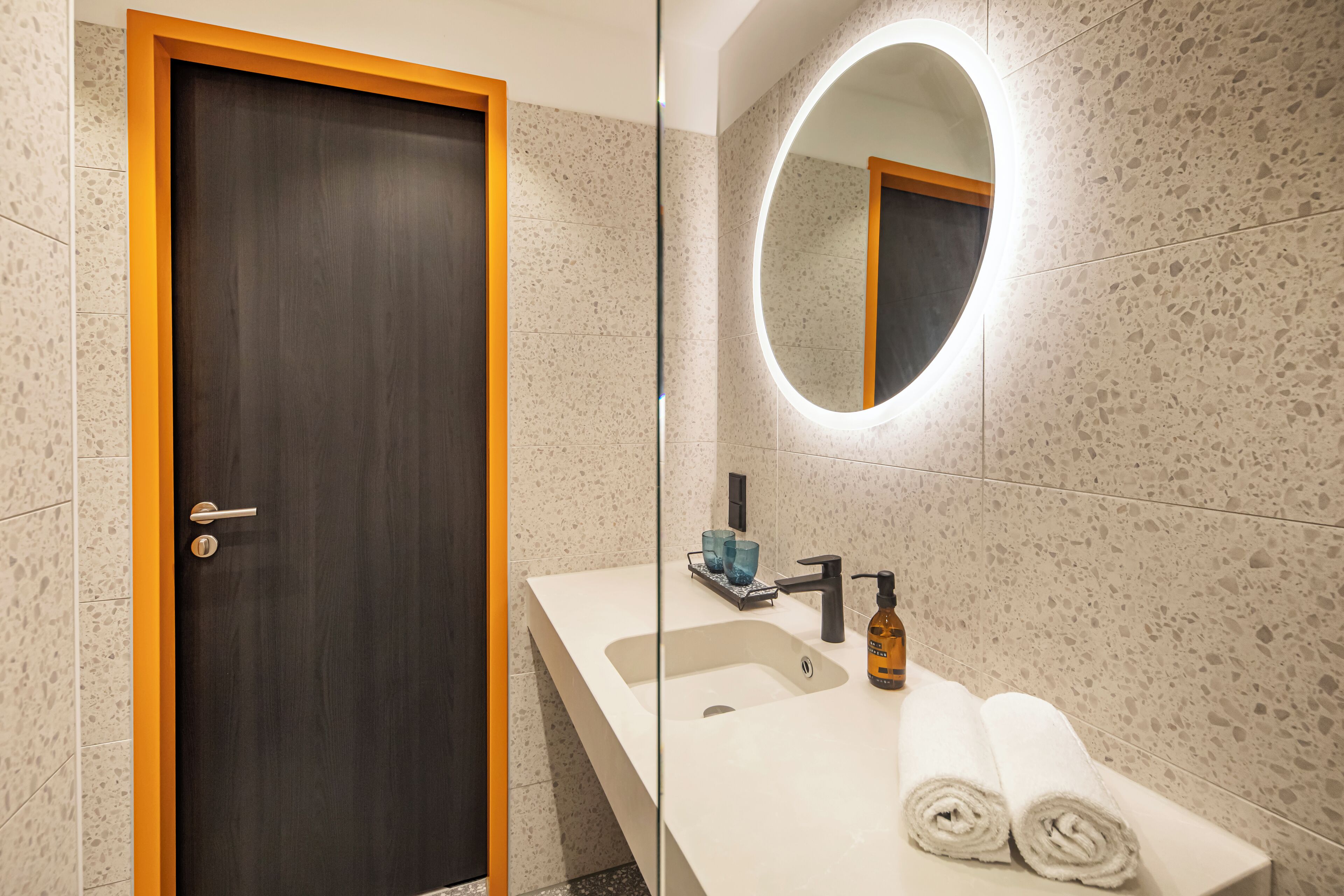 A modern bathroom with a dark wooden door, a white sink with a round mirror, and two rolled towels on the counter.