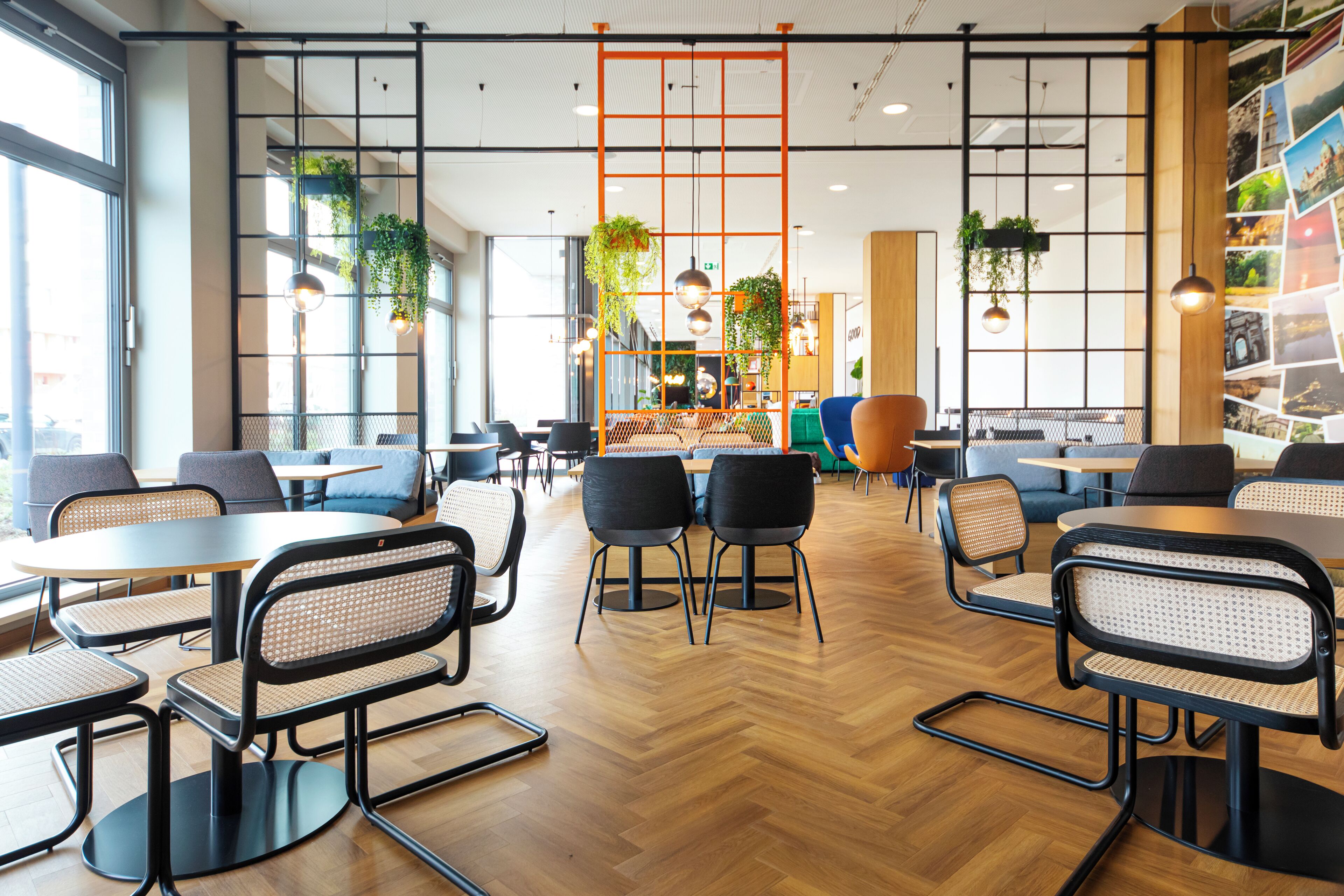Ein modernes Café mit Holzböden, schwarzen und grauen Stühlen sowie Tischen. Hängende Pflanzen und eine große, orangefarbene Gitterstruktur befinden sich in der Mitte.