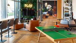 Een moderne loungehoek met een tafeltennistafel, comfortabele zithoek, een neonbord met 'PLAY' en een wand versierd met planten en foto's.