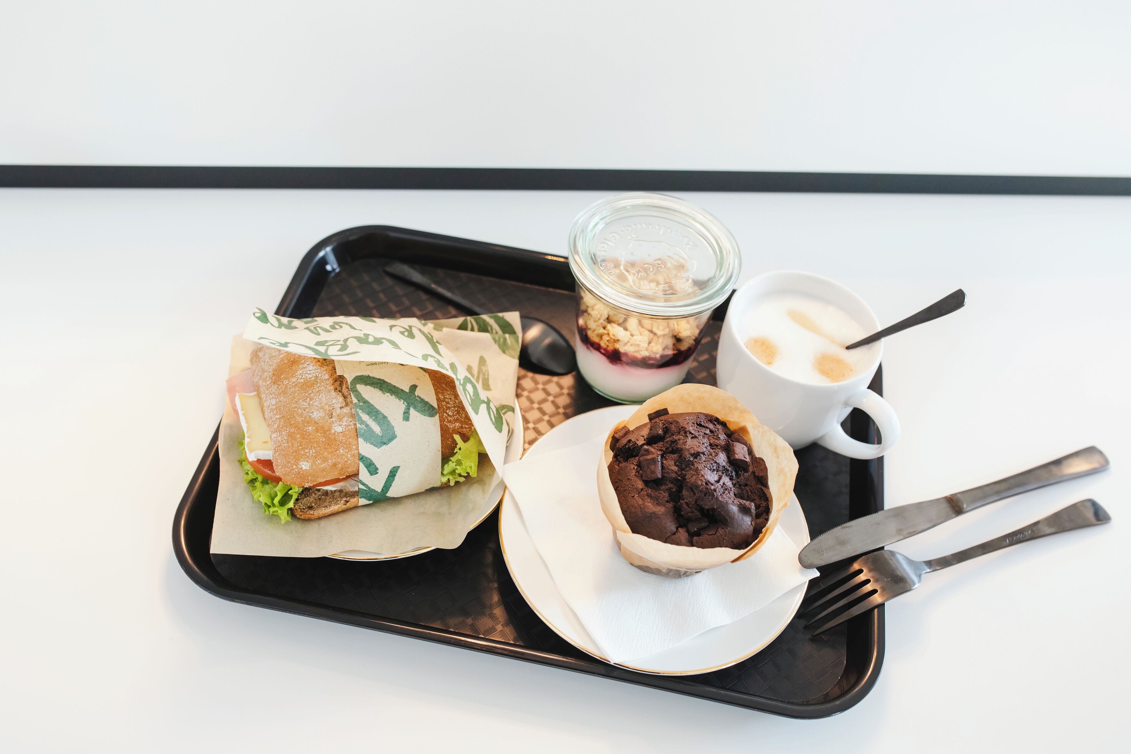 Uma bandeja com sanduíche, muffin, xícara de café e copo de parfait de iogurte.