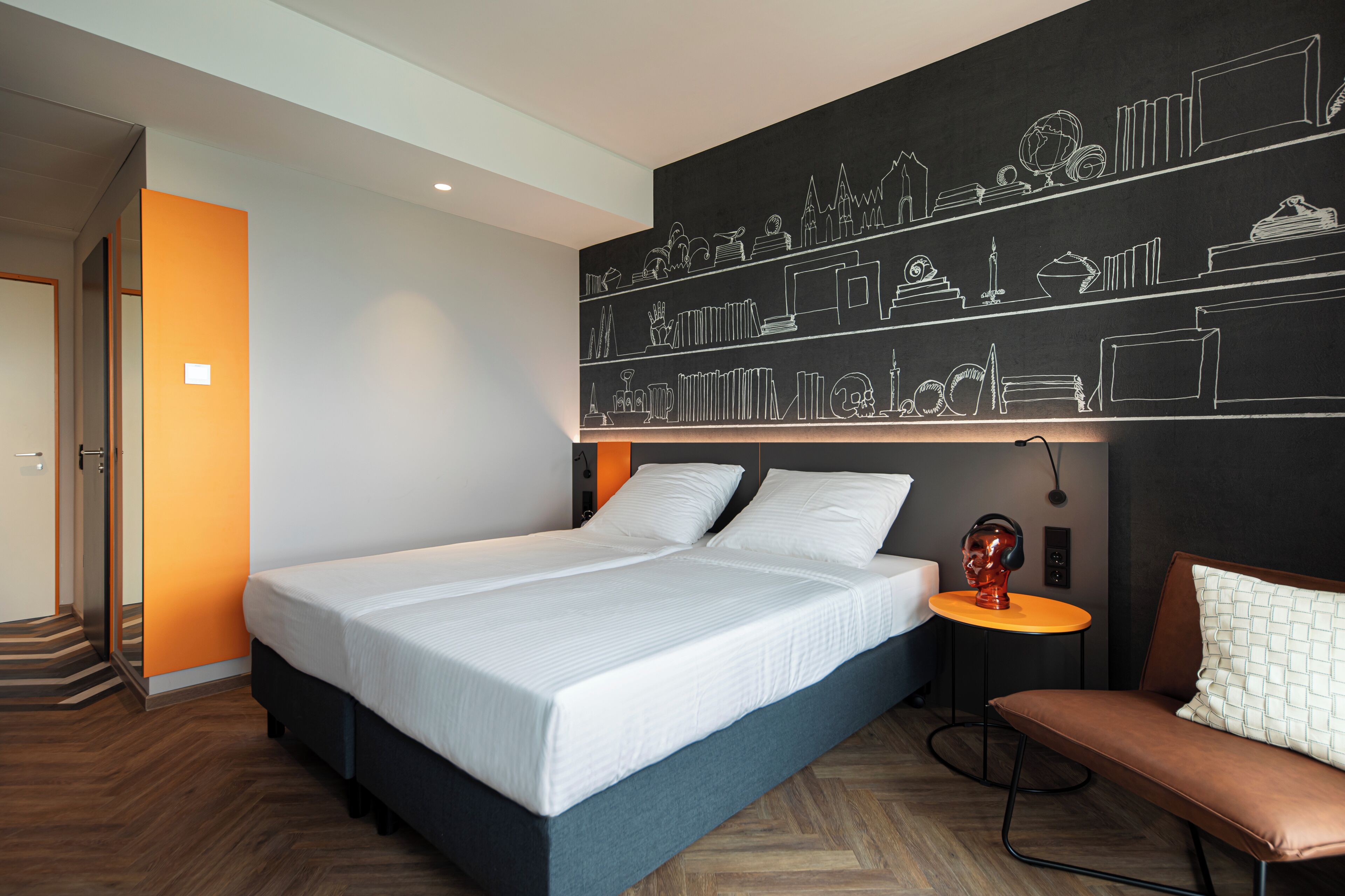 Una habitación de hotel moderna con una cama grande, una mesita de noche con una lámpara, un banco y una pared de pizarra con varios dibujos.