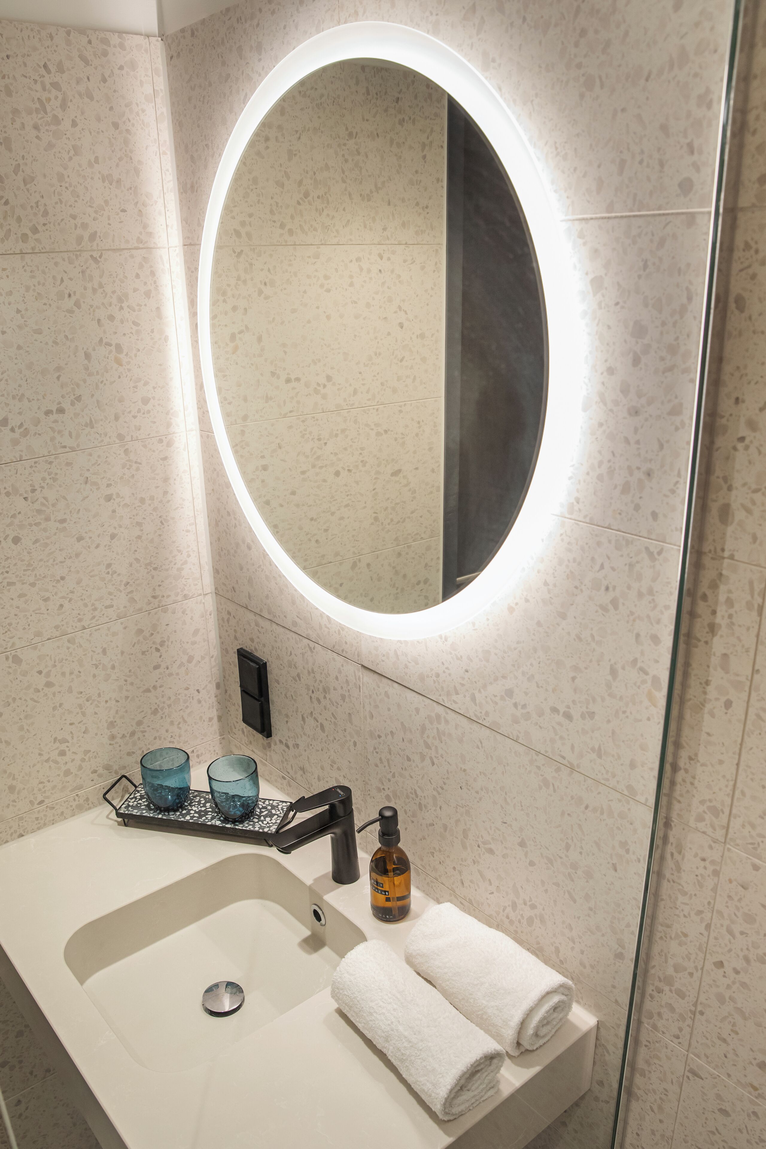 A bathroom with a white sink, a round mirror with a light, and two blue glasses on a tray.