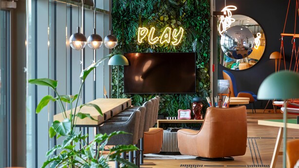 Een moderne lounge met een neonbord waarop 'PLAY' staat, een grote flatscreen-tv en comfortabele zitplaatsen.
