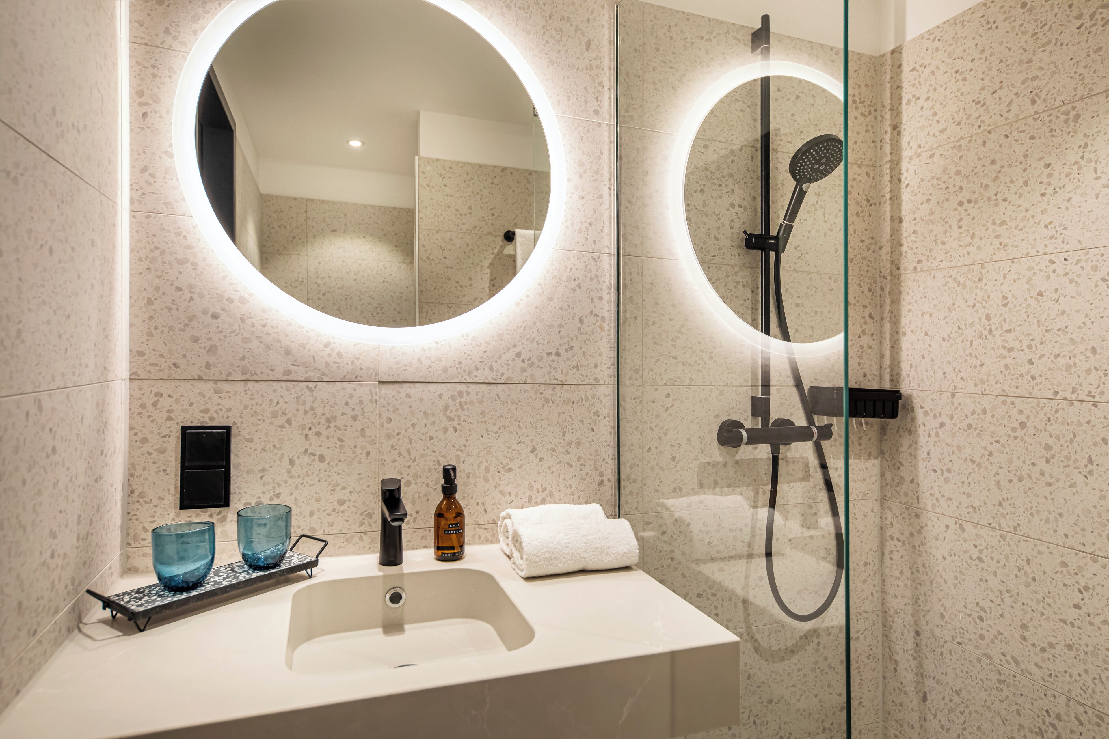 A modern bathroom with a round mirror, a glass-enclosed shower, and a white sink with a tray holding two blue glass bowls and a bottle.