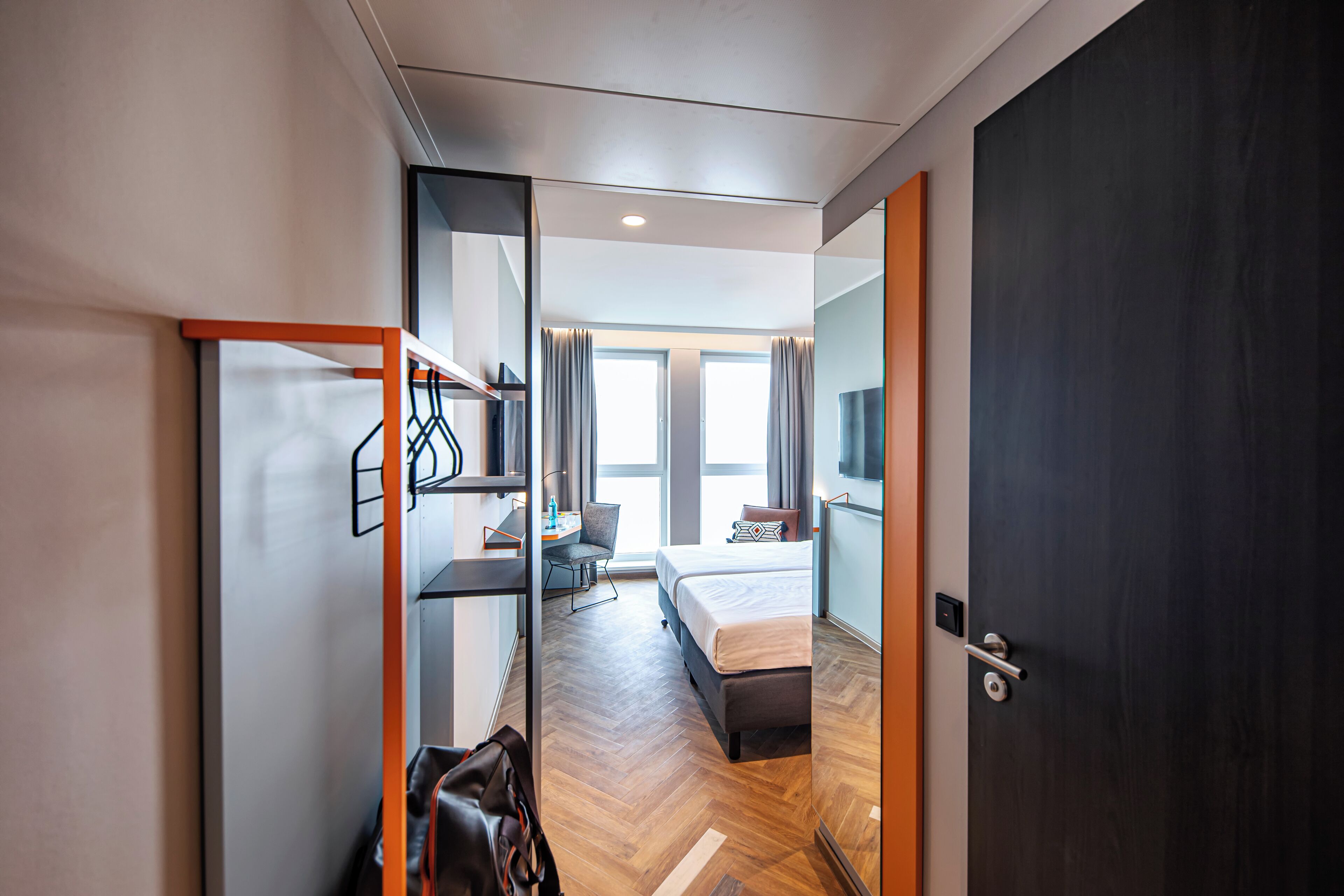 A modern hotel room with a bed, a desk, a chair, and a wardrobe.