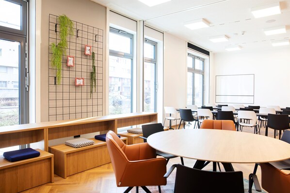 A modern conference room with a round table, comfortable seating, a grid wall with plants, and large windows. - LOGINN Hotel Köln Airport (Cologne)