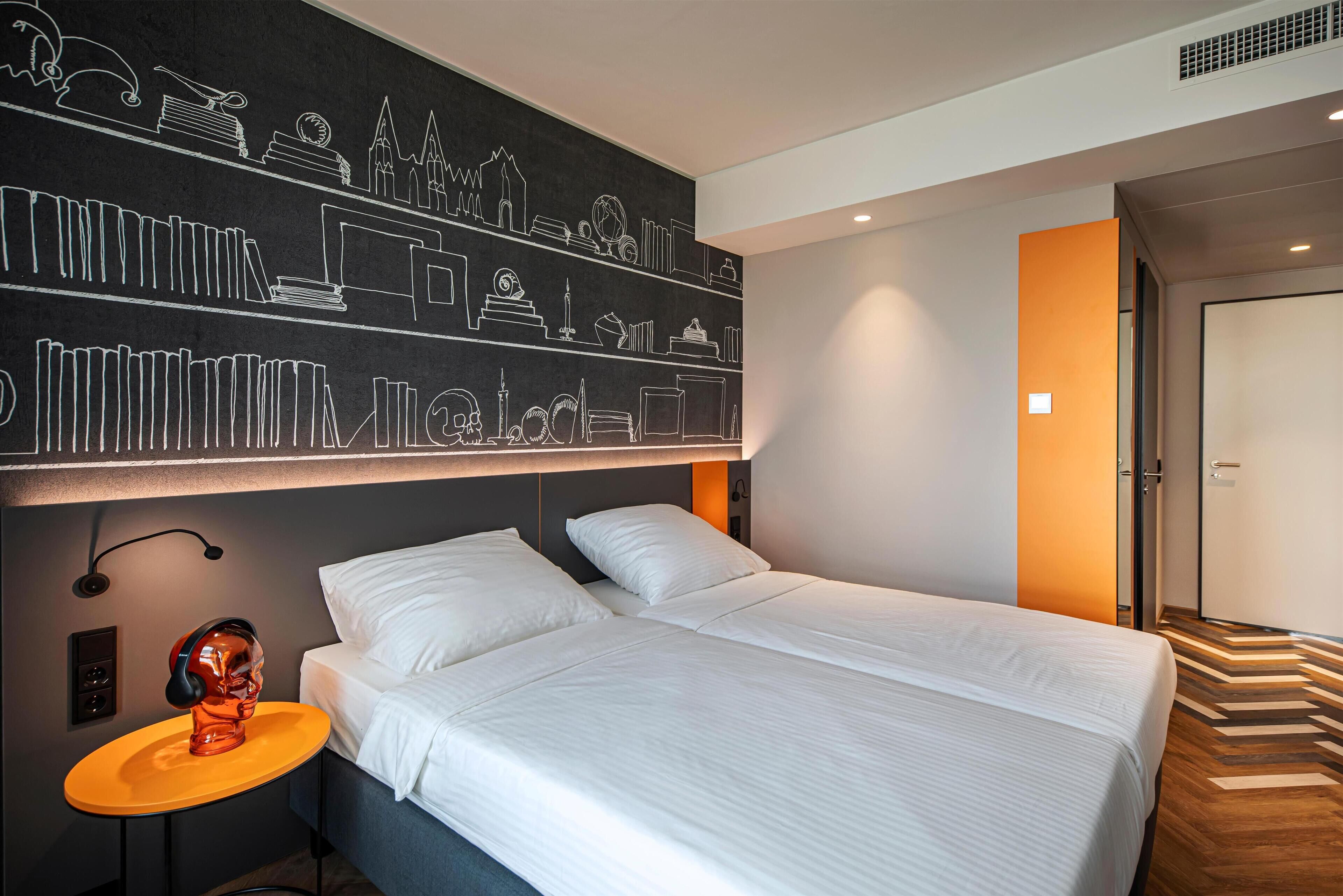 A modern hotel room with a large bed, a bedside table with a skull-shaped lamp, and a wall decorated with cityscape drawings.