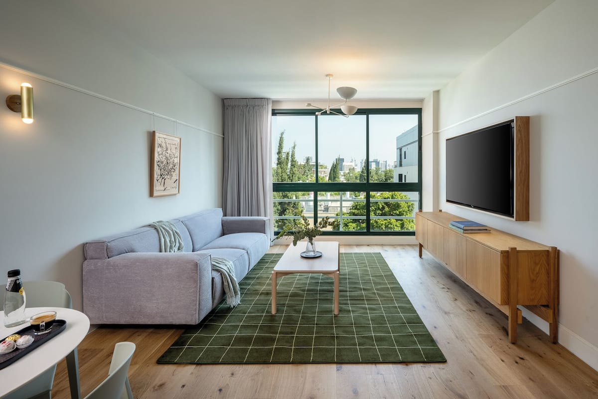 Un salon moderne avec un canapé, une petite table, un téléviseur fixé au mur et une grande fenêtre donnant sur la ville.