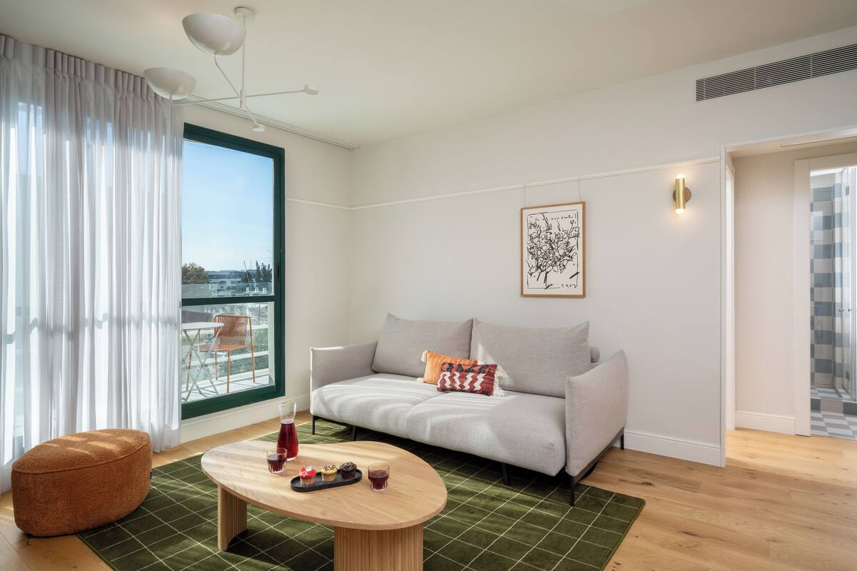 Un salon moderne avec un canapé, une table basse ronde, un tapis vert et une vue dégagée sur l’extérieur grâce à une grande fenêtre.