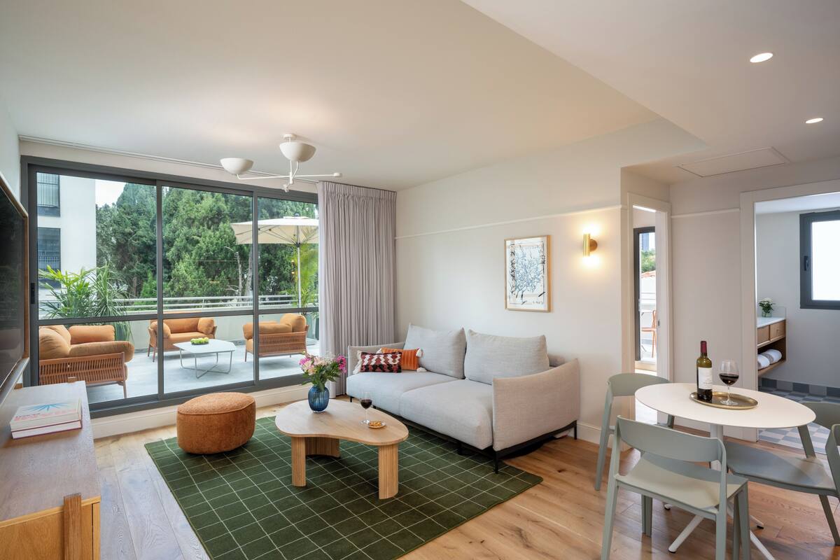 Un salon moderne avec un canapé, une table basse et une table à manger. Une grande fenêtre offre une vue sur la verdure environnante.
