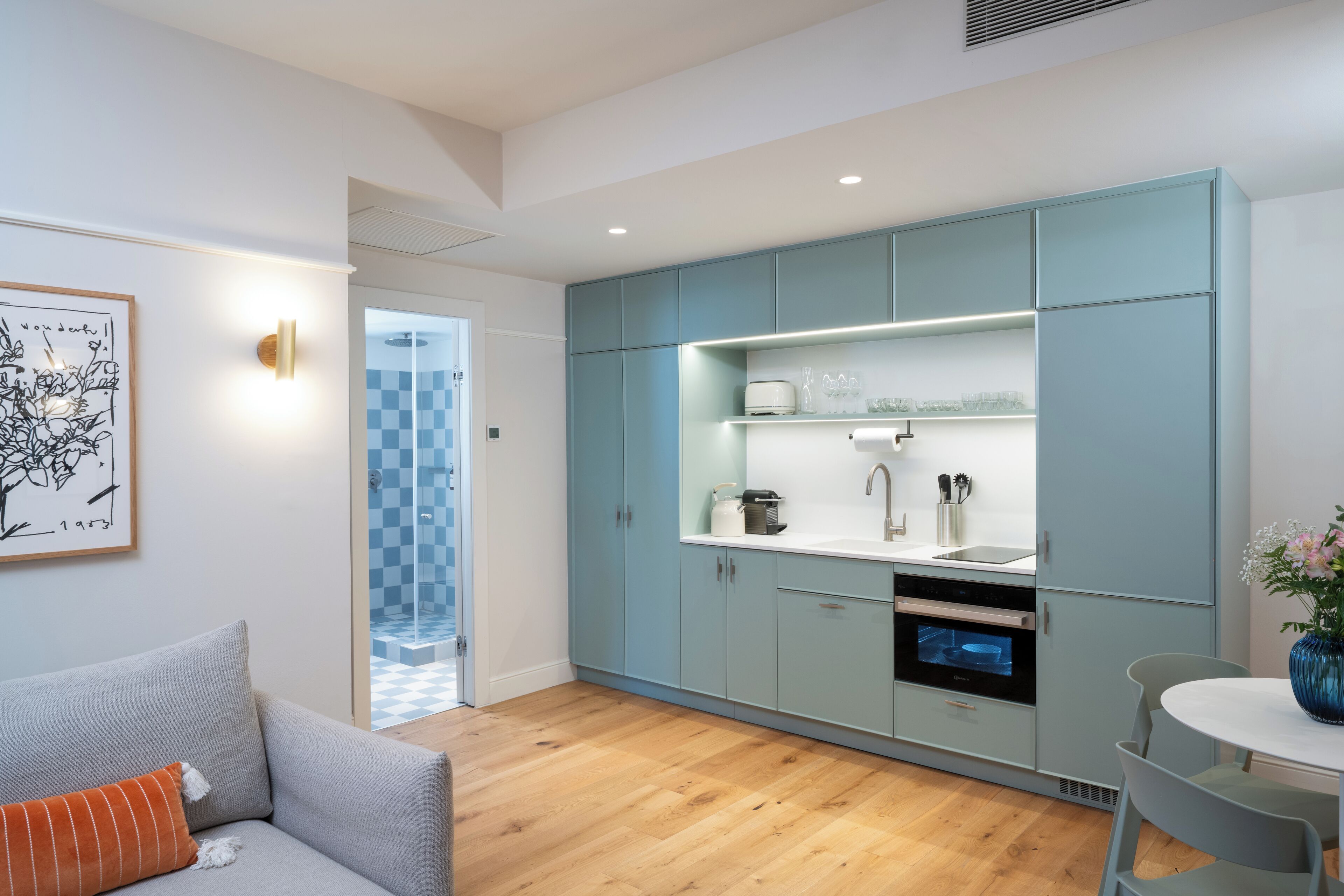 A modern kitchen with light blue cabinets, a white countertop, and a built-in oven. There is a grey sofa with an orange pillow in the adjacent living area.
