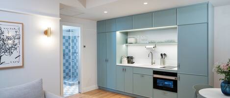 A modern kitchen with light blue cabinets, a white countertop, and a built-in oven. There is a grey sofa with an orange pillow in the adjacent living area.