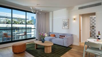 Ein modernes Wohnzimmer mit Sofa, Couchtisch und Hocker, großen Fenstern mit Blick auf Gebäude und Grünflächen, sowie einem Essbereich mit Stühlen und einem Tisch.