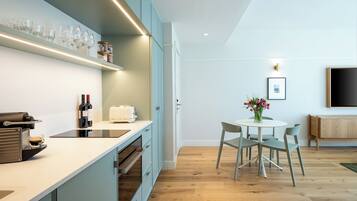 A modern kitchen with a built-in oven, a coffee machine, and wine bottles. There is a dining area with a table and chairs, and a TV mounted on the wall.