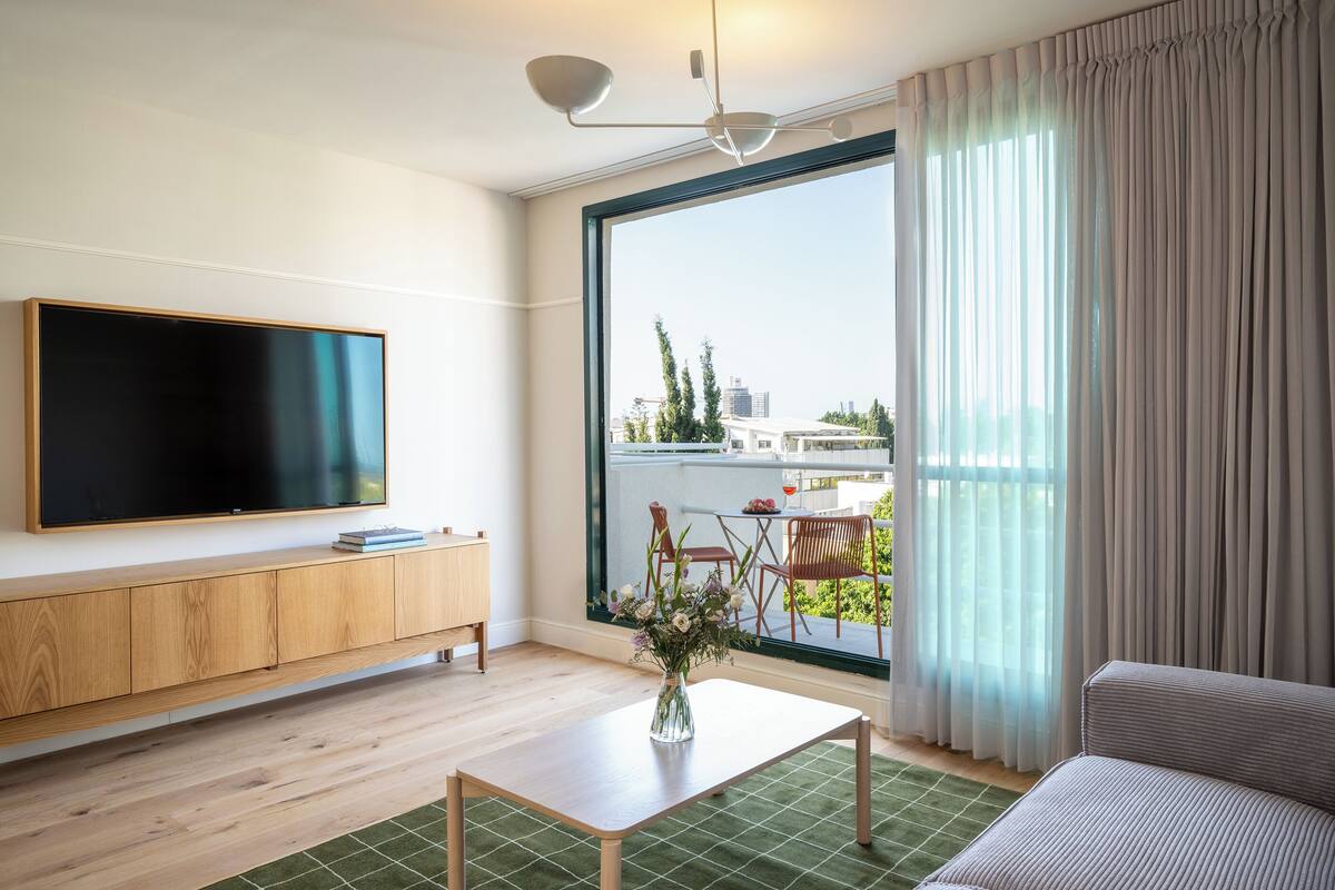Un salon moderne doté d’un téléviseur à écran plat, de meubles en bois, d’un tapis vert et offrant une vue sur l’extérieur grâce à de grandes fenêtres.