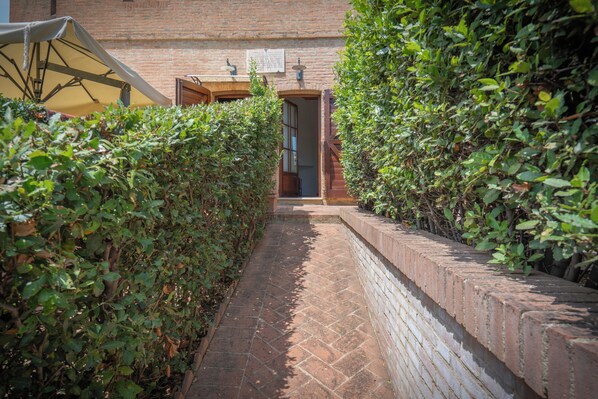 Un estrecho sendero de ladrillo bordeado por setos verdes frondosos, que conduce a un edificio con un acceso abierto.