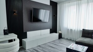 A modern living room with a flat-screen TV mounted on a dark wall, a white sofa, a coffee table with a board game, and a white chair. - Loft & Living am Geiseltalsee (Braunsbedra)