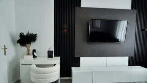 A modern living room with a flat-screen TV mounted on a dark wall, a white desk with a chair, and a decorative plant. - Loft & Living am Geiseltalsee (Braunsbedra)