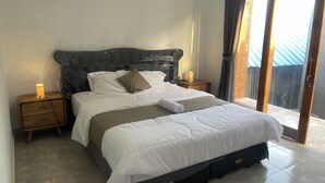 A neatly made bed with white bedding and a folded towel, a wooden nightstand with a lamp, and a view of a building through a sliding glass door. - Zee House (Canggu)