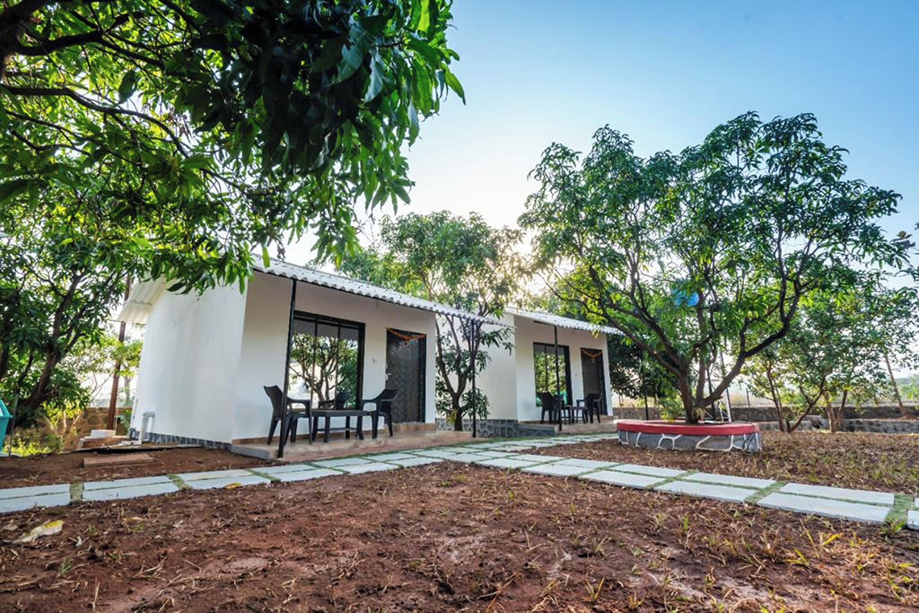 Un bâtiment à deux étages, à l’extérieur blanc, doté d’un patio couvert avec des sièges extérieurs, et d’un jardin avec des arbres et un sentier en pierre.