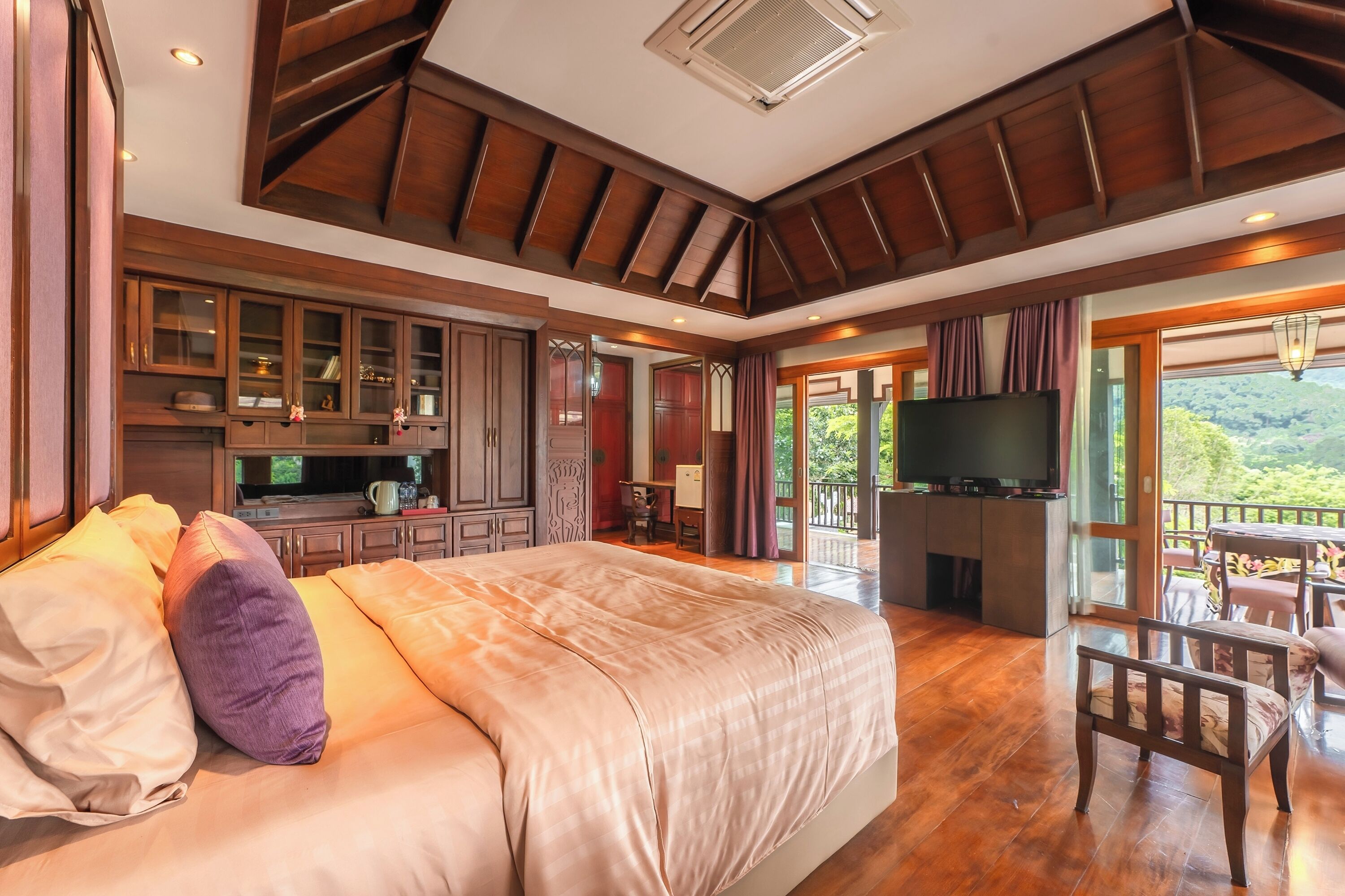 A spacious bedroom with a large bed, wooden ceiling beams, a TV, and a balcony with a scenic view.