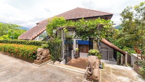 Una casa con tetto in tegole, ingresso con cancello e giardino con fiori rossi e due statue di elefanti.