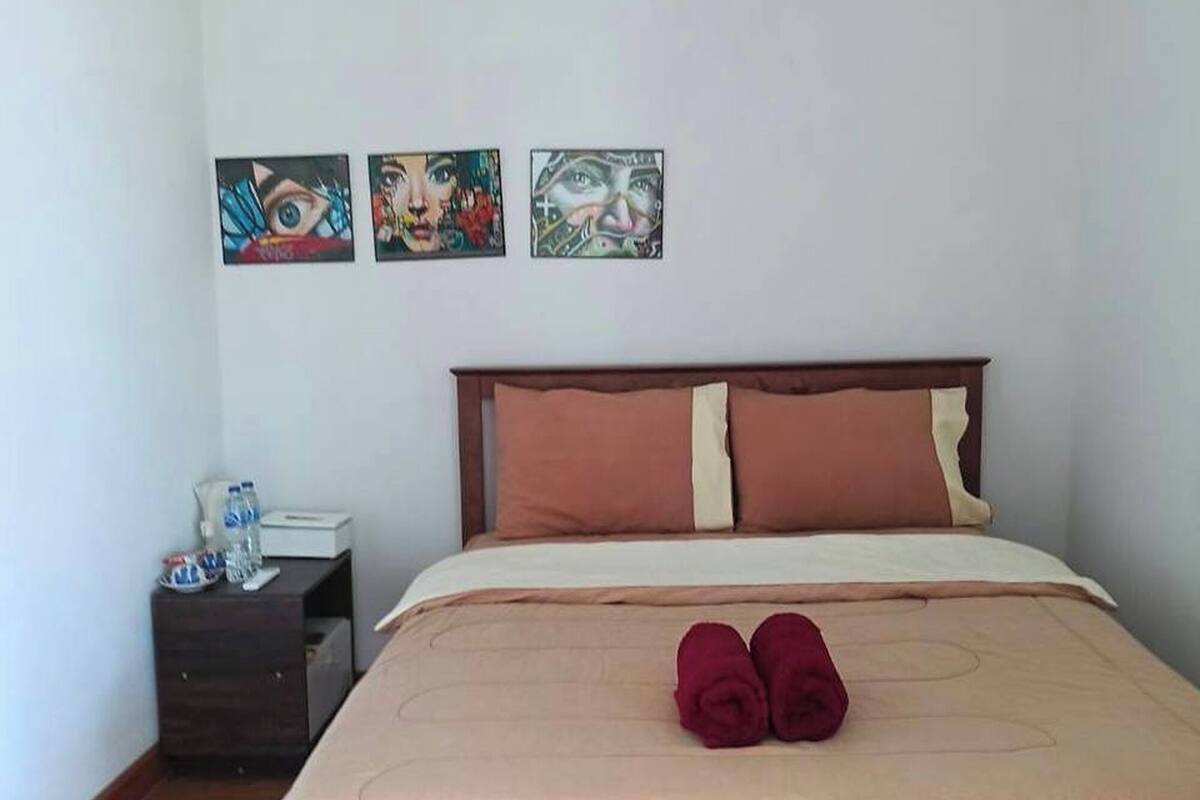 Un lit bien fait, recouvert d’une couvre-lit beige et agrémenté de deux oreillers, une table de chevet en bois avec une bouteille d’eau et une boîte de mouchoirs, et trois œuvres d’art accrochées au mur.