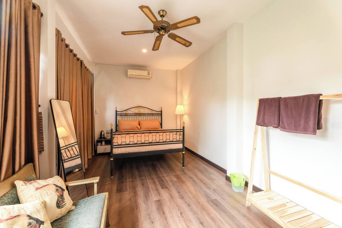 Une chambre à coucher comprenant un lit, un ventilateur de plafond, une table de chevet, une lampe, une chaise et une petite étagère en bois.